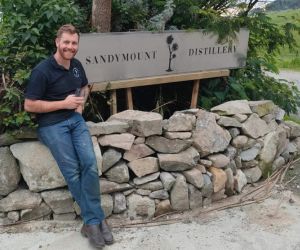 Richard Wilson sitting on a stone wall at the gate of Sandymount Distillery, with the silver sign behind him.